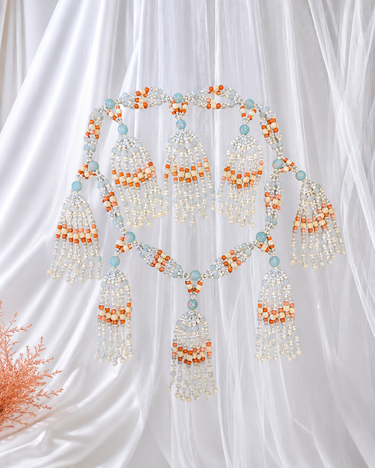 An  ornate beaded necklace with tassels  in white, bone, opal and coral colored  beads,  arranged against a white draped fabric background.