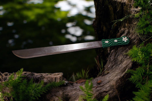 A machete with a beaded handle, featuring cowrie shells and green and black decorative beads, near  a tree trunk.