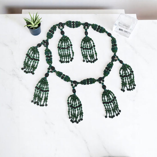 Green and black beaded necklace on a white surface with a small plant.