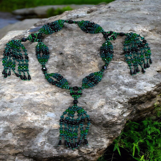 A beaded necklace set with green and black colors, featuring a detailed pattern and ornamental designs, displayed on a natural stone.
