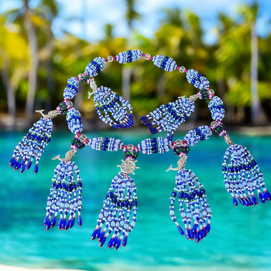 A beaded mazo featuring a combination of blue, green, white, opal, and pink coral colors, with accents of a ship's wheel and mermaids, displayed against a tropical beach backdrop.