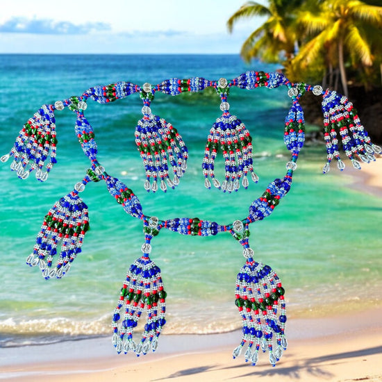 A beaded mazo necklace featuring a variety of colors including blue, green, white, pink coral, and transparent, displayed against a beach backdrop with palm trees and ocean in the background.