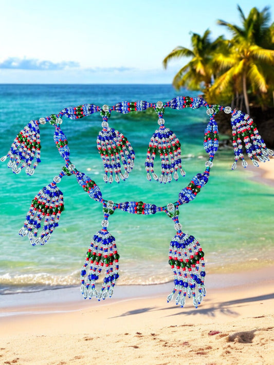 A beaded mazo necklace featuring a variety of colors including blue, green, white, pink coral, and transparent, displayed against a beach backdrop with palm trees and ocean in the background.