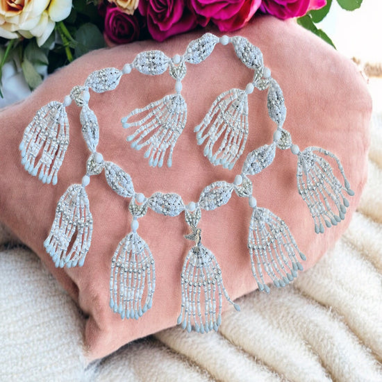 A beaded necklace with white, pearl, and mother of pearl strings arranged in a symmetrical pattern with dove adornments, displayed on a pink coral cushion.