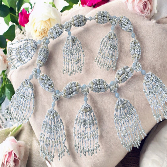 A detailed image of a 10-string Oshanla-inspired mazo, featuring intricate beadwork with white, pearl, and mother of pearl accents, displayed on a soft fabric surface with floral arrangements in the background.