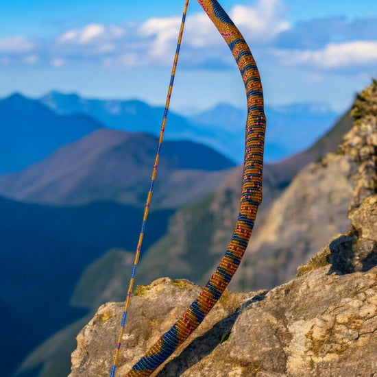 A handcrafted glass beaded bow with blue, coral, and topaz beads, displayed on a rock with a mountainous landscape in the background.