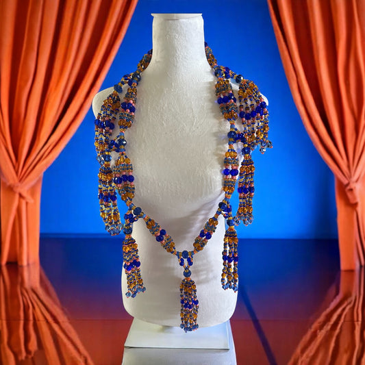 A mannequin displaying a Yoruba mazo necklace with blue and amber Czech glass beads against an orange curtain backdrop.