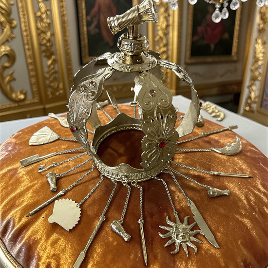 Decorative gold metal crown with adornments on a amber velvet pillow with ornate background.