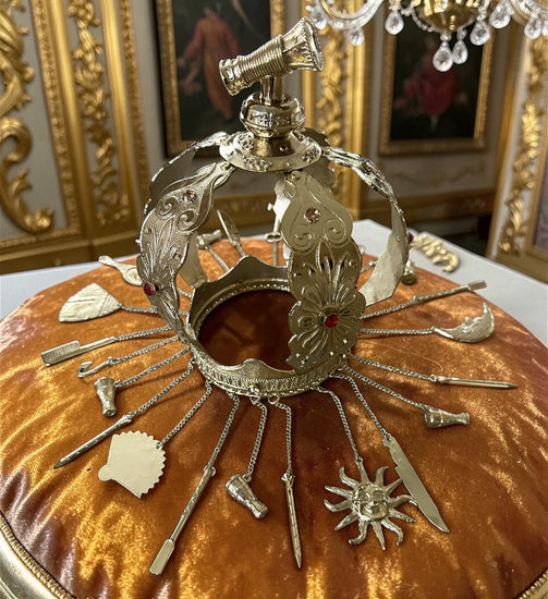 Decorative gold metal crown with adornments on a amber velvet pillow with ornate background.