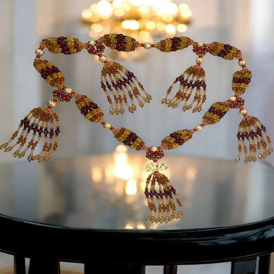 A detailed image of a glass beaded mazo, featuring multiple strings with beads in various colors including gold, red, and black, arranged in a fan-like structure, displayed on a reflective surface.