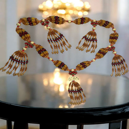 A detailed image of a glass beaded mazo, featuring multiple strings with beads in various colors including gold, red, and black, arranged in a fan-like structure, displayed on a reflective surface.