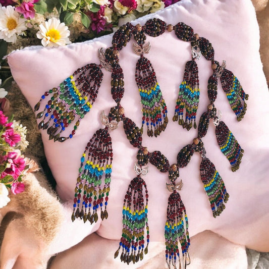 A detailed image of a traditional African-style beaded necklace with multiple strands and copper masquerade masks, displayed on a cushion with a floral background.