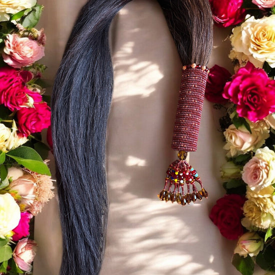 A beaded horse tail with a handle adorned with brown and multicolored beads, and copper accents, presented on a white surface surrounded by an array of colorful flowers.