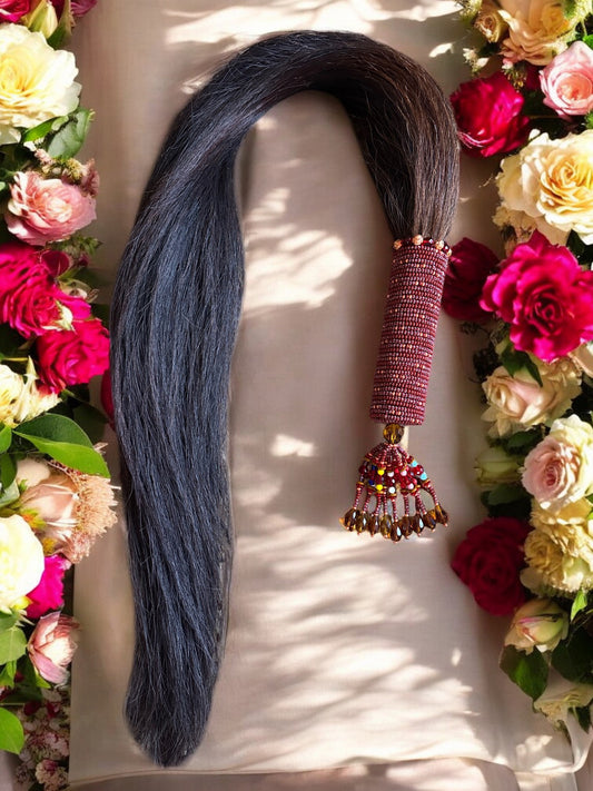 A beaded horse tail with a handle adorned with brown and multicolored beads, and copper accents, presented on a white surface surrounded by an array of colorful flowers.