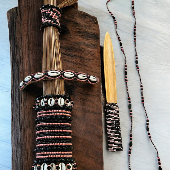 A set of traditional Yoruba spiritual cleansing items including a beaded cleansing artifact, a bamboo obé, and a traditional necklace, displayed on a wooden board.