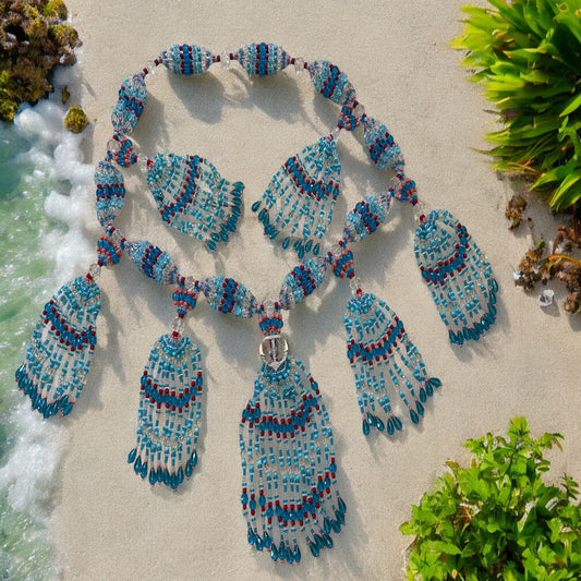 A beaded glass mazo featuring aqua blue, light blue, red, transparent, and pink coral tones, laid out on a sandy beach with ocean waves and greenery in the background.