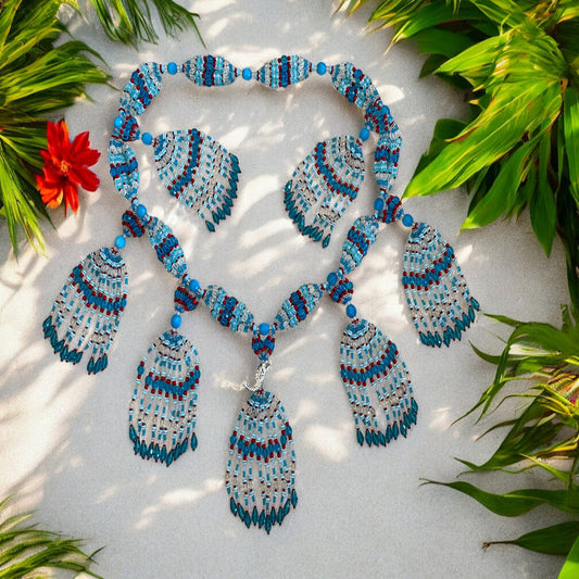 A beaded glass mazo featuring a mermaid charm, with colors including blue, pink, and red, displayed on a white surface surrounded by green foliage.