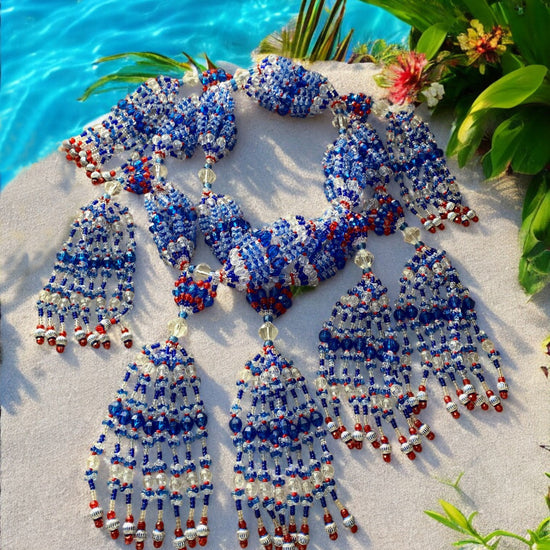 A beaded necklace set laid out on a sandy surface with a representation of Yemayá, featuring blue, red, and clear beads with silver accents, along with a body of water and greenery in the background.