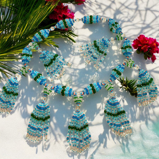 A beaded glass mazo with a color palette of aqua blue, light blue, green, transparent, and pink coral, displayed on a white surface with a shadow of a plant and vibrant red flowers in the background.