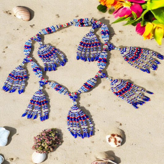 A beaded necklace with blue, red, and transparent strings laid out on a sandy surface, surrounded by seashells and flowers.