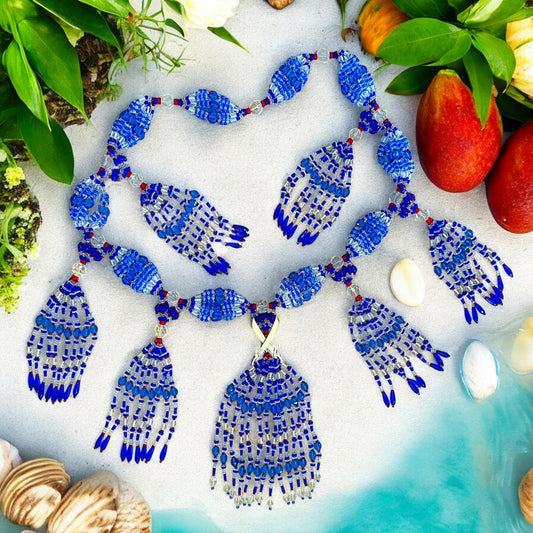 A beaded necklace with a geometric pattern, featuring sapphire blue, transparent, and bold red colors, laid out on a surface with shells and fruits and adorned with cross machete charm.