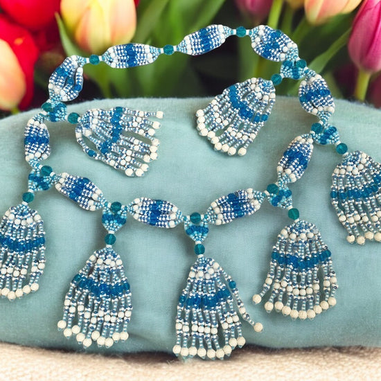 A beaded necklace with white, light blue, and aquamarine strings arranged in a symmetrical pattern with tassel-like ends, displayed against a floral background.