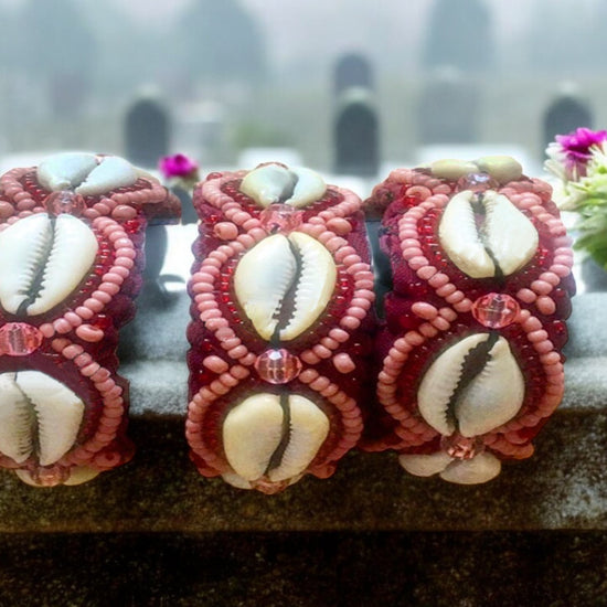 Three handcrafted bracelets with light pink and translucent red beads, adorned with cowrie shells, displayed against a floral backdrop.