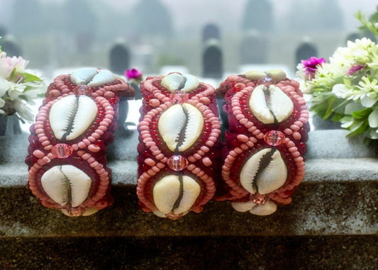 Three handcrafted bracelets with light pink and translucent red beads, adorned with cowrie shells, displayed against a floral backdrop.